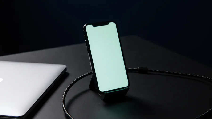 A phone in a stand on a dark desk, cabled to a laptop